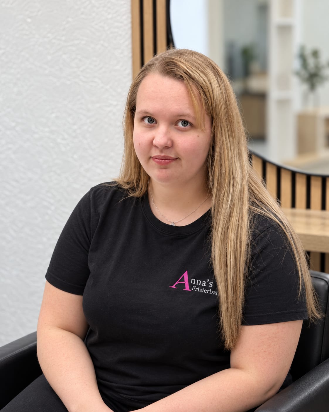 Anna-Lena Reents im schwarzen Shirt mit dem Logo von Anna’s Frisierbar, hinter ihr ein runder Spiegel an den Holzlamellen