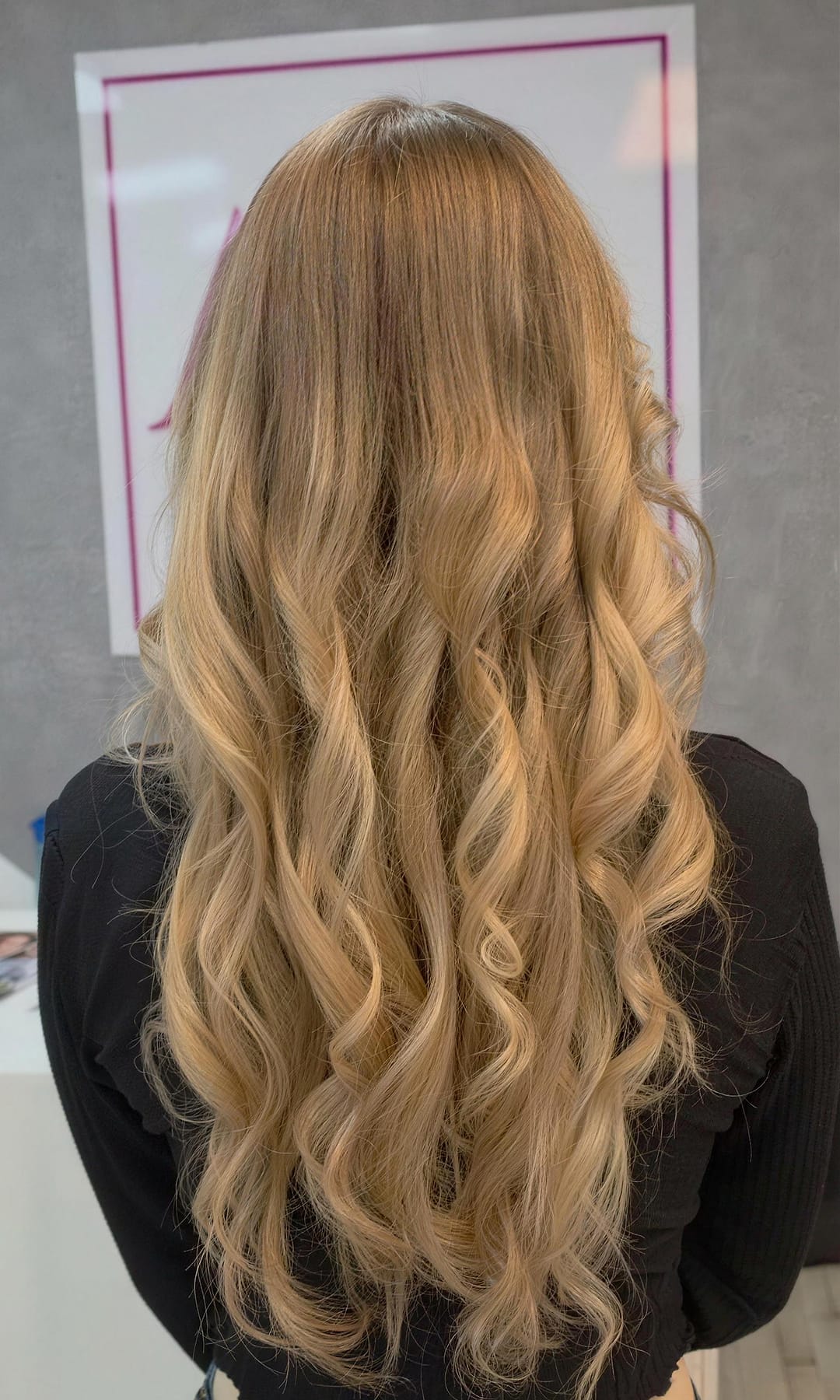 Lange, gewellte Haare in Karamell- und Honigblond, von hinten fotografiert, vor dem Logo-Schild im Salon